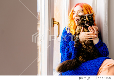 Emotional portrait woman with orange hair is holding a cat in her arms. The cat is black and white. The woman is smiling and she is happy 115538500