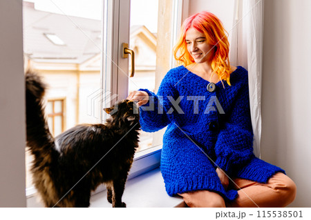 A woman with orange hair is petting a cat while wearing a blue cozy sweater. The cat is sitting on a windowsill. Pet lover concept. 115538501