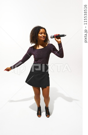 Young woman, singer in fashionable attire singing in microphone isolated on white background. Music and Fashion Young woman, singer in fashionable attire singing in microphone isolated on white background. Music and Fashion 115538516