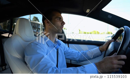 Profile of handsome businessman driving a modern electric car at urban road. Male successful entrepreneur riding on electrical vehicle at city. Young man commuting to work at sunny day. Slow motion 115539103