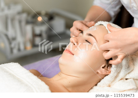 Young woman enjoying facial massage, lying on spa bed indoor at beauty salon. 115539473