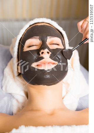 Cosmetologist applying black sheet mask on woman's face in beauty salon. Cosmetologist applying black sheet mask on woman's face in beauty salon. 115539507