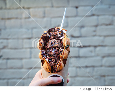 Hong Kong waffles in female hand on brick wall background. Delicious bubble waffle with chocolate biscuit cookies and chocolate syrup in woman hand. Trendy food. Hong Kong waffles in female hand on brick wall background. Delicious bubble waffle with chocolate biscuit cookies and chocolate syrup in woman hand. Trendy food. 115541699