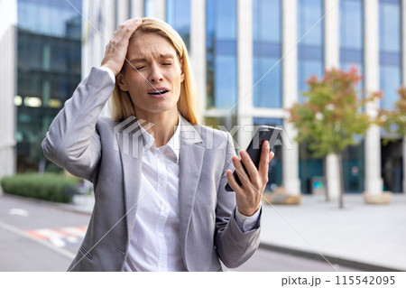 Worried and upset young business woman standing outside on the street in a suit, holding a mobile phone and holding her head with her hand in disappointment, got bad news. 115542095