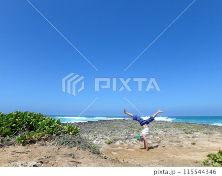 Man does split leg Handstand on coral rocks on the beach as wave crash in the distance Man does split leg Handstand on coral rocks on the beach as wave crash in the distance 115544346