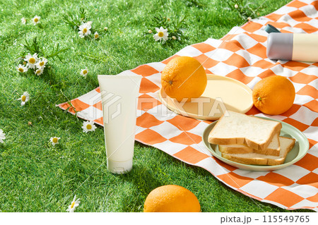 Advertising photo for beauty care products with outdoor picnic concept, a white cosmetic tube without label placed on the green lawn, beside an orange rug contains some oranges and sandwiches above 115549765