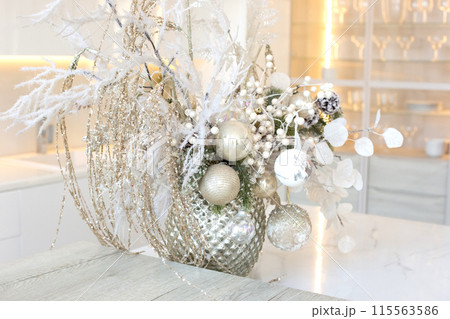 A silver vase with white branches and balls stands on a table in a white kitchen interior A silver vase with white branches and balls stands on a table in a white kitchen interior 115563586