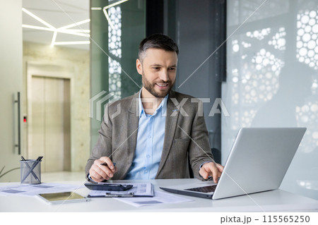 Smiling businessman working on laptop in modern office, analyzing documents and taking notes. Professional and focused in corporate environment. Smiling businessman working on laptop in modern office, analyzing documents and taking notes. Professional and focused in corporate environment. 115565250