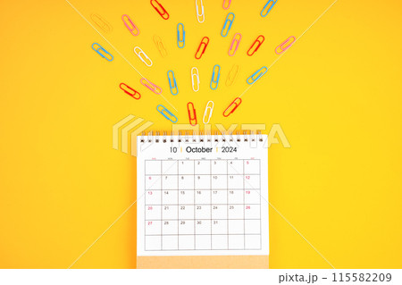 October 2024 desk calendar and colorful metal paperclips on yellow background. 115582209