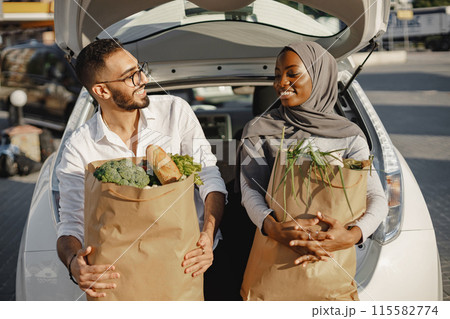 Happy diverse couple having fun after shopping groceries. Sitting on the car trunk with full of fresh and healthy food. African muslim woman and arab man. Happy diverse couple having fun after shopping groceries. Sitting on the car trunk with full of fresh and healthy food. African muslim woman and arab man. 115582774