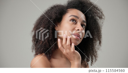 Beauty and healthcare concept - beautiful African American woman with curly afro hairstyle and clean, healthy skin touches her cheek and face with her hand, posing and looking at the camera 115584560