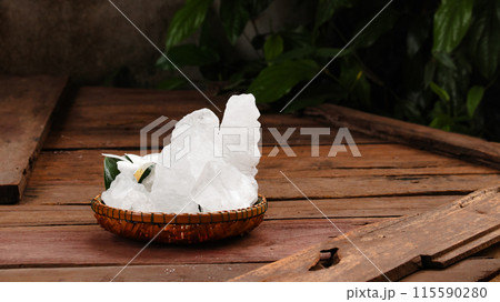 A basket filled with white alum crystals sits on a vibrant green shrub background. 115590280