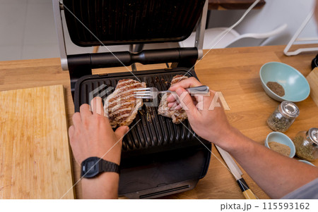 Chef at the kitchen preparing beef steaks on the home electric grill 115593162