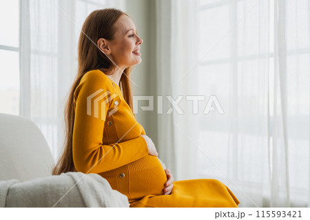 Pregnancy motherhood people expectation future. Pregnant woman with big belly sitting on chair near window at home. Girl hugging her tummy enjoying pregnancy. Maternity tenderness parenthood new life 115593421