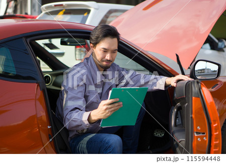 A mechanic in work uniform with a document clipboard, smile while inspect a car coming for maintenance at a repair shop. A mechanic in work uniform with a document clipboard, smile while inspect a car coming for maintenance at a repair shop. 115594448