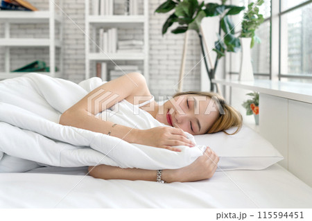 Young asian woman in white undershirt, sleep and sweet dream lying on bed in a bright, cozy room with pillow and a comforter. Side view Young asian woman in white undershirt, sleep and sweet dream lying on bed in a bright, cozy room with pillow and a comforter. Side view 115594451