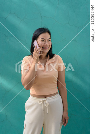 Asian woman against a turquoise wall wearing a light peach polo shirt using mobile phone Asian woman against a turquoise wall wearing a light peach polo shirt using mobile phone 115595444