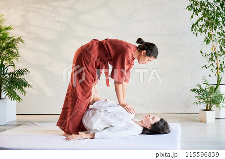 Masseuse making thai yoga massage. Woman therapist doing traditional massage treatment, stretching female patient's body. Client lying on mat, enjoying hand massage in calm atmosphere. Masseuse making thai yoga massage. Woman therapist doing traditional massage treatment, stretching female patient's body. Client lying on mat, enjoying hand massage in calm atmosphere. 115596839