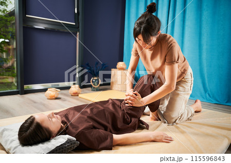 Masseuse making thai yoga massage. Woman therapist doing traditional massage treatment for female patient indoors. Client lying on mat, meditating, enjoying hand massage in calm atmosphere. Masseuse making thai yoga massage. Woman therapist doing traditional massage treatment for female patient indoors. Client lying on mat, meditating, enjoying hand massage in calm atmosphere. 115596843
