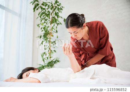 Masseuse making thai yoga massage. Woman therapist doing traditional massage treatment for female patient using elbow. Client lying on mat, meditating, enjoying hand massage in calm atmosphere. 115596852
