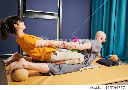 Masseuse making thai yoga massage. Woman therapist doing traditional massage treatment, stretching male patient's body. Bald client lying on mat, enjoying hand massage in calm atmosphere. Masseuse making thai yoga massage. Woman therapist doing traditional massage treatment, stretching male patient's body. Bald client lying on mat, enjoying hand massage in calm atmosphere. 115596857