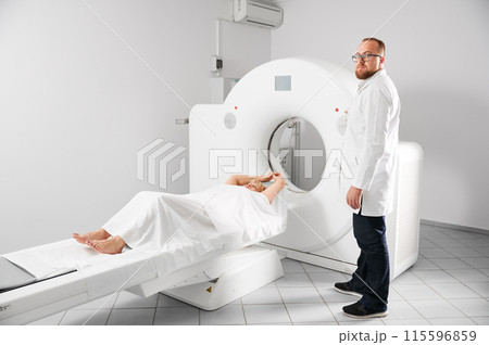 Medical computed tomography or MRI scanner. Male radiologist presses MRI button to examine female patient, looking to the camera. Concept of healthcare and modern diagnostics. Medical computed tomography or MRI scanner. Male radiologist presses MRI button to examine female patient, looking to the camera. Concept of healthcare and modern diagnostics. 115596859