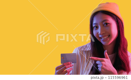 Banking service. Money transaction. Pink neon light happy hipster girl customer recommending credit card electronic payment isolated on yellow empty space background. Banking service. Money transaction. Pink neon light happy hipster girl customer recommending credit card electronic payment isolated on yellow empty space background. 115596961