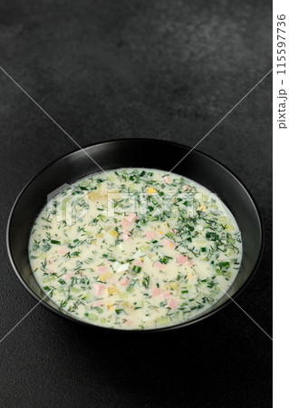 Cold soup with fresh cucumbers, sausage, broth and sour cream in a black plate on a black background. A traditional dish is okroshka Cold soup with fresh cucumbers, sausage, broth and sour cream in a black plate on a black background. A traditional dish is okroshka 115597736