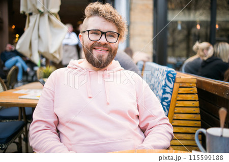 Young bearded man having breakfast at table of street cafe on spring day, drinking warm cappuccino and enjoy morning. 115599188