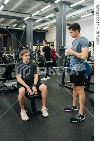 Full length vertical shot of muscular personal instructor explaining technique of exercise to young beginner sportsman sitting on bench with barbell during fitness training. Concept of healthy life. Full length vertical shot of muscular personal instructor explaining technique of exercise to young beginner sportsman sitting on bench with barbell during fitness training. Concept of healthy life. 115599395