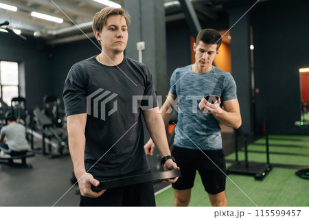 Dedicated beginner sportsman holding and prepare to rotating weight disc plate overhead during sport workout training in modern gym, support under supervision of personal trainer. Dedicated beginner sportsman holding and prepare to rotating weight disc plate overhead during sport workout training in modern gym, support under supervision of personal trainer. 115599457