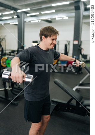 Vertical shot of motivated beginner sportsman doing side lateral raises with dumbbells at gym. Active handsome man exercising to stay fit and slim at sport club. Concept of healthy sports life. Vertical shot of motivated beginner sportsman doing side lateral raises with dumbbells at gym. Active handsome man exercising to stay fit and slim at sport club. Concept of healthy sports life. 115599482