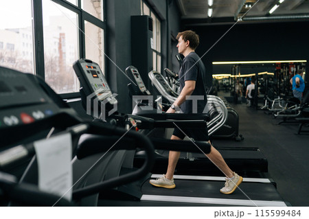 Full length side shot of athletic man walking on treadmill preforming warming-up run routine exercise in gym. Sporty male walking on running track, working on health and fitness at sport club. 115599484
