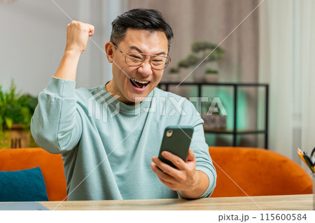Asian Chinese man at home use smartphone celebrating success victory winning play online game Asian Chinese man at home use smartphone celebrating success victory winning play online game 115600584