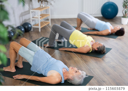 Elderly woman in company of hatha yoga lovers performs bridge pose, setu bandha sarvangasana Elderly woman in company of hatha yoga lovers performs bridge pose, setu bandha sarvangasana 115603879