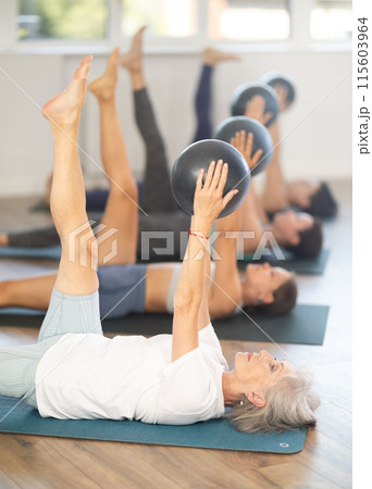 Senior woman performing leg lifts with exercise ball in group Pilates class 115603964