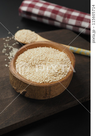 A wooden bowl filled with white sesame seeds, accompanied by a spoon, set on a dark surface 115605714