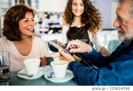 Elderly couple paying for their coffe with debit card. University student working part-time in cafe. Elderly couple paying for their coffe with debit card. University student working part-time in cafe. 115614598