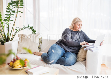 Overweight woman working on laptop from homeoffice, sitting on sofa, making video call. Overweight woman working on laptop from homeoffice, sitting on sofa, making video call. 115614783