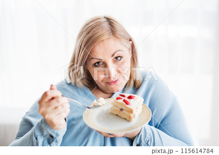 Portrait of beautiful young overweight woman at home, eating slice of cake, enjoying sweet taste. Portrait of beautiful young overweight woman at home, eating slice of cake, enjoying sweet taste. 115614784