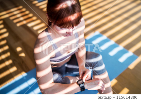 Beautiful redhead woman checking her performance on smartwatch after home fitness routine. Home workout on gym mt in living room, sporty woman wearing sportswear. 115614880