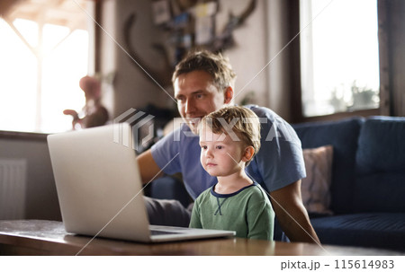 Dad and boy watching cartoons on laptop, sitting on sofa in living room, having great time together. Dad and boy watching cartoons on laptop, sitting on sofa in living room, having great time together. 115614983