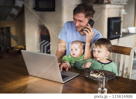 Father sitting at table working on laptop, two sons sitting by him. Older brother eating porridge. Unconditional parental love, family moment. 115614986