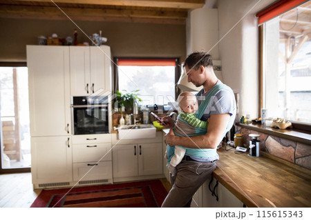 Father holding small baby n baby carrier, standing in kitchen scrolling on smartphone. Unconditional paternal love, father's Day. 115615343