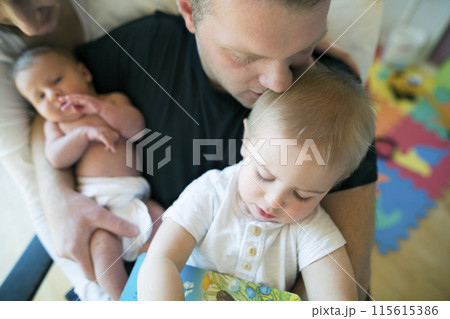 Father holding newborn baby daughter, while reading book with older son. Dad spending time with kids in room. Unconditional paternal love, Father's Day. 115615386
