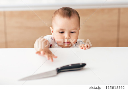 A curious baby, dressed in a white outfit with colorful patterns, reaches for a knife placed on a kitchen counter. The background features wooden cabinets and a light-colored wall A curious baby, dressed in a white outfit with colorful patterns, reaches for a knife placed on a kitchen counter. The background features wooden cabinets and a light-colored wall 115618362
