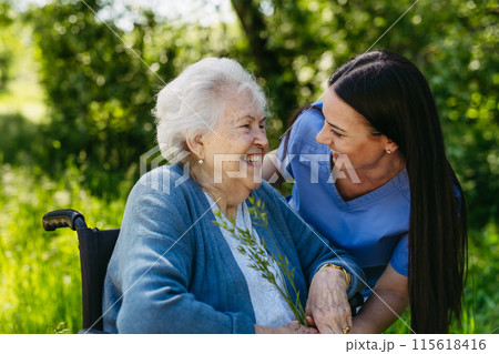 Female caregiver and senior woman in wheelchair picking wild flowers. Nurse and elderly woman enjoying a warm day in nursing home, public park. Female caregiver and senior woman in wheelchair picking wild flowers. Nurse and elderly woman enjoying a warm day in nursing home, public park. 115618416