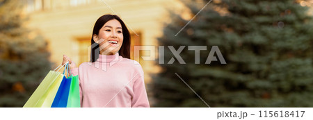 Asian woman is standing outdoors holding multiple shopping bags in her hands. She appears to have just finished shopping and is carrying her purchases. The bags are colorful and seem full, panorama 115618417