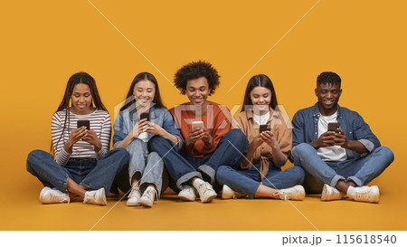 This image features a diverse group of five young adults, all smiling and looking down at their smartphones. They are sitting cross-legged on a yellow background This image features a diverse group of five young adults, all smiling and looking down at their smartphones. They are sitting cross-legged on a yellow background 115618540