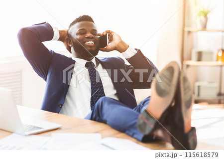 Positive african american manager talking by phone while having break at office, leaning back on chair and putting legs on table, empty space Positive african american manager talking by phone while having break at office, leaning back on chair and putting legs on table, empty space 115618579
